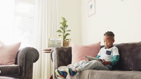 Happy-african-american-boy-sitting-on-sofa-using-tablet,-in-slow-motion