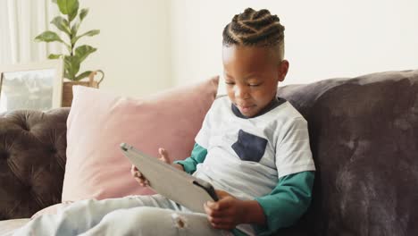 Glücklicher-Afroamerikanischer-Junge,-Der-Auf-Dem-Sofa-Sitzt-Und-Ein-Tablet-Benutzt,-In-Zeitlupe