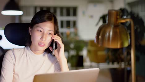 Mujer-De-Negocios-Asiática-Casual-Hablando-Por-Teléfono-Inteligente-Y-Usando-Una-Computadora-Portátil-En-La-Oficina-Por-La-Noche,-Cámara-Lenta