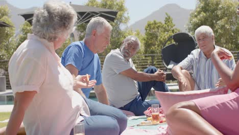 Happy-diverse-senior-friends-having-picnic-and-talking-in-sunny-garden,-unaltered,-in-slow-motion