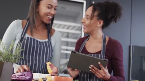 Happy-biracial-lesbian-couple-in-aprons-preparing-food-and-using-tablet-in-kitchen,-in-slow-motion