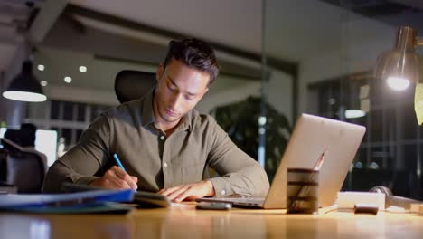 Vielbeschäftigter-Gemischtrassiger-Geschäftsmann-Am-Schreibtisch-Mit-Laptop-Und-Notizen-Im-Büro-Nachts,-Zeitlupe