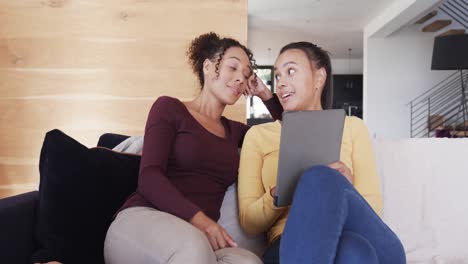 Happy-biracial-lesbian-couple-relaxing-on-couch-together,-using-tablet-and-talking,-in-slow-motion