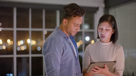 Focused-diverse-male-and-female-colleague-standing-talking-and-using-tablet-at-night-in-office