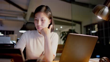 Asian-businesswoman-sitting-at-desk,-using-laptop-and-tablet-at-office,-in-slow-motion