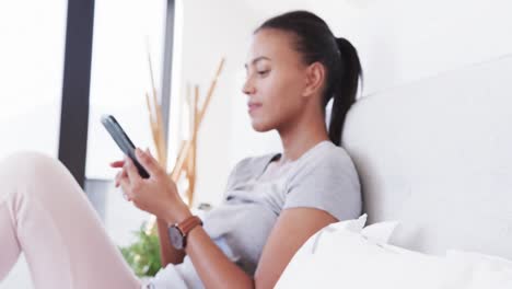 Happy-biracial-woman-sitting-on-bed-using-smartphone,-in-slow-motion
