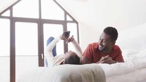 Happy-african-american-couple-lying-on-bed-using-smartphone-and-talking-at-home,-in-slow-motion