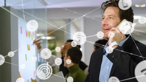 Animation-of-network-of-digital-icons-over-caucasian-businessman-talking-on-smartphone-at-office