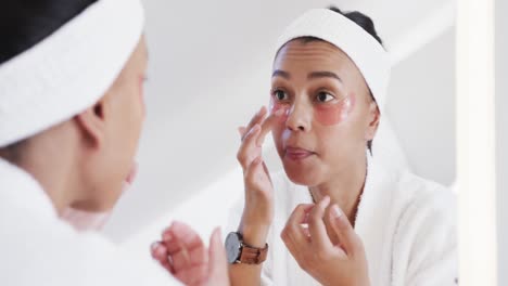 Focused-biracial-woman-applying-under-eye-masks-looking-in-mirror-in-sunny-bathroom,-slow-motion