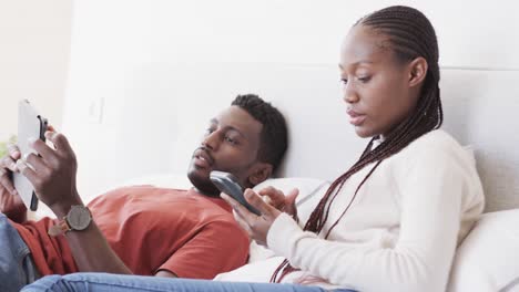African-american-couple-lying-on-bed-using-smartphone-and-tablet,-talking-at-home,-in-slow-motion