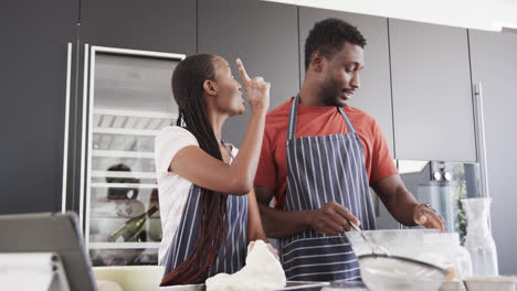 Happy-african-american-couple-in-aprons-baking-together-using-tablet-in-kitchen,-slow-motion