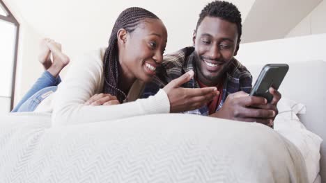 Glückliches-Afroamerikanisches-Paar,-Das-Zu-Hause-Auf-Dem-Bett-Liegt-Und-In-Zeitlupe-Auf-Das-Smartphone-Schaut