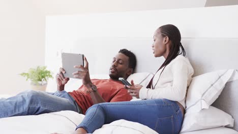 Glückliches-Afroamerikanisches-Paar,-Das-Auf-Dem-Bett-Liegt-Und-In-Zeitlupe-Mit-Smartphone-Und-Tablet-Spricht