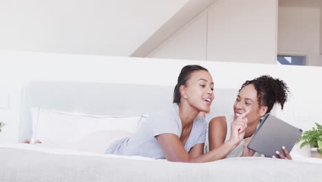 Happy-biracial-lesbian-couple-lying-on-bed-using-tablet-together,-in-slow-motion