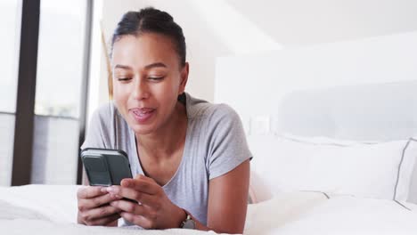 Happy-biracial-woman-lying-on-bed-using-smartphone-and-smiling,-in-slow-motion