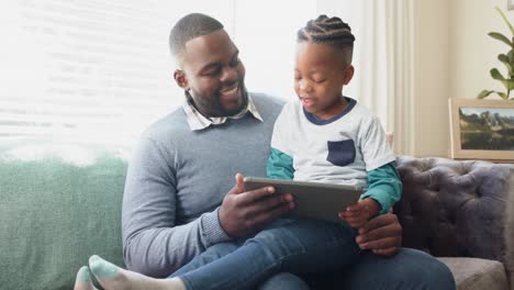 Glücklicher-Afroamerikanischer-Vater,-Der-Mit-Seinem-Sohn-Auf-Dem-Schoß-Auf-Dem-Sofa-Sitzt-Und-Ein-Tablet-Benutzt,-In-Zeitlupe