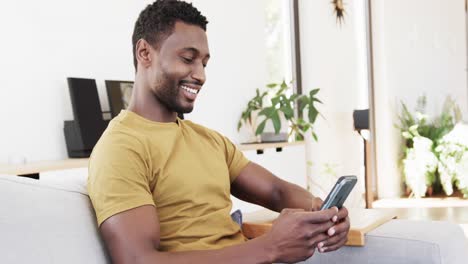 Glücklicher-Afroamerikanischer-Mann-Mit-Smartphone,-Der-Auf-Der-Couch-Im-Wohnzimmer-Sitzt,-In-Zeitlupe