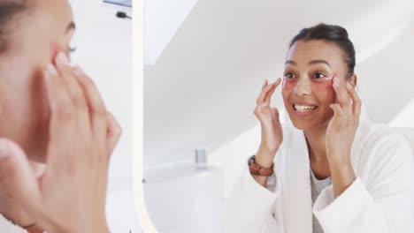 Happy-biracial-woman-applying-under-eye-masks-looking-in-mirror-in-sunny-bathroom,-slow-motion