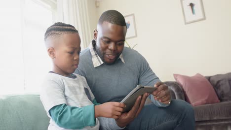 Happy-african-american-father-and-son-sitting-on-sofa-using-tablet-in-living-room,-in-slow-motion