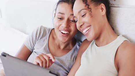 Happy-biracial-lesbian-couple-sitting-on-bed-using-tablet,-talking-and-embracing,-in-slow-motion