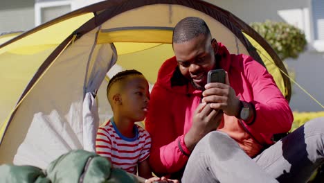 Happy-african-american-father-and-son-in-tent-using-smartphone-in-sunny-garden,-in-slow-motion