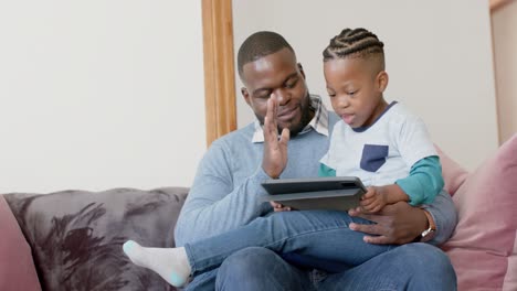 Happy-african-american-father-and-son-sitting-on-sofa-using-tablet-and-high-fiving,-in-slow-motion