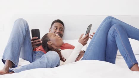 Glückliches-Afroamerikanisches-Paar,-Das-Auf-Dem-Bett-Liegt-Und-In-Zeitlupe-Redet-Und-Smartphones-Benutzt