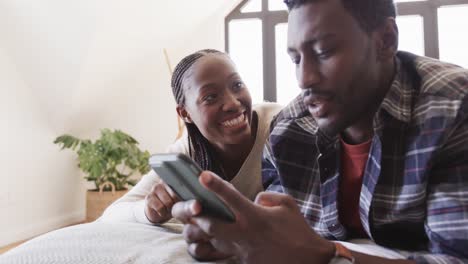 Glückliches-Afroamerikanisches-Paar,-Das-Mit-Dem-Smartphone-Im-Bett-Liegt-Und-Zu-Hause-Redet,-In-Zeitlupe