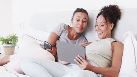 Happy-biracial-lesbian-couple-sitting-on-bed-using-tablet-and-smartphone-and-talking,-in-slow-motion