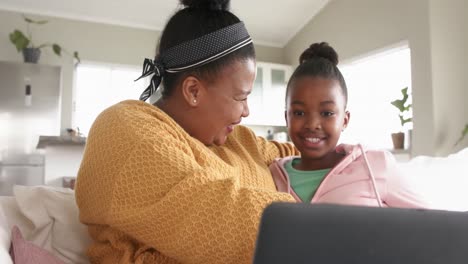 Happy-african-american-mother-and-daughter-relaxing-on-sofa-using-laptop-and-hugging,-in-slow-motion