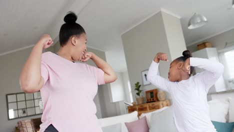 Happy-unaltered-african-american-mother-and-daughter-flexing-muscles-in-living-room,-in-slow-motion