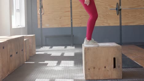 Low-section-of-unaltered-biracial-woman-jumping-on-box,-cross-training-at-gym,-slow-motion