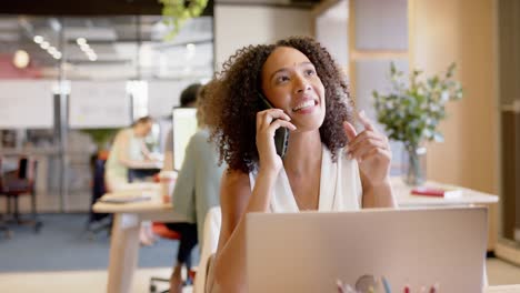 Mujer-De-Negocios-Birracial-Hablando-Por-Teléfono-Inteligente-Y-Usando-Una-Computadora-Portátil-En-La-Oficina,-En-Cámara-Lenta