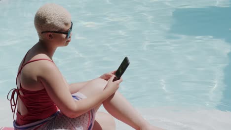 Happy-biracial-woman-using-smartphone-and-sitting-at-swimming-pool-in-slow-motion