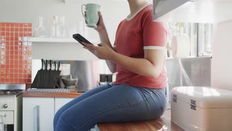 Happy-biracial-woman-drinking-coffee-and-using-smartphone-at-home-in-slow-motion
