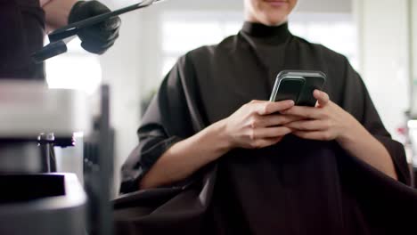 Midsection-of-caucasian-female-client-having-hair-dyed-using-smartphone-at-salon,-in-slow-motion
