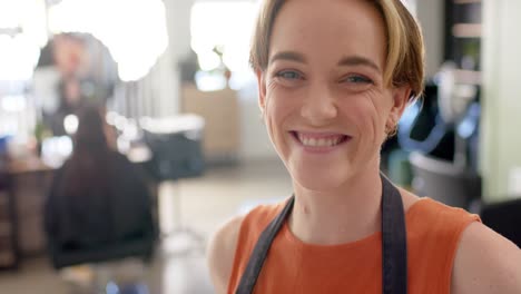 Portrait-of-happy-caucasian-female-hairdresser-in-apron-smiling-at-hair-salon,-slow-motion