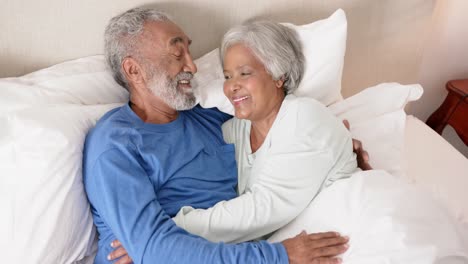 Happy-senior-biracial-couple-lying-on-bed,-embracing-and-smiling,-unaltered,-in-slow-motion