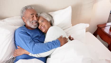 Happy-senior-biracial-couple-lying-on-bed-and-embracing,-unaltered,-in-slow-motion