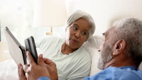 Glückliches-älteres-Gemischtrassiges-Paar,-Das-Auf-Dem-Bett-Liegt-Und-Tablet-Und-Smartphone-Benutzt,-Unverändert,-In-Zeitlupe