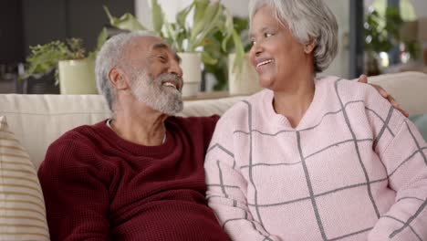 Portrait-of-happy-senior-biracial-couple-sitting-on-sofa-and-embracing-,-unaltered,-in-slow-motion