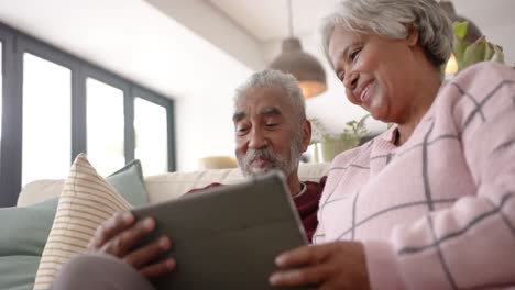 Glückliches-älteres-Gemischtrassiges-Paar,-Das-Auf-Dem-Sofa-Sitzt-Und-Ein-Tablet-Benutzt,-Unverändert,-In-Zeitlupe