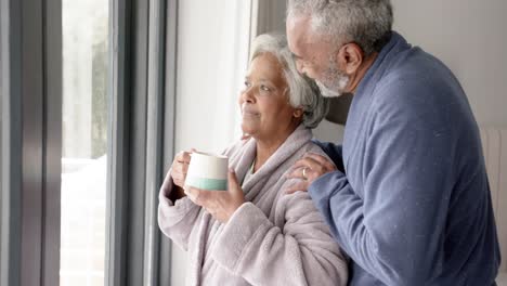 Glückliches-älteres-Gemischtrassiges-Paar,-Das-Durch-Das-Fenster-Schaut-Und-Sich-In-Zeitlupe-Umarmt,-Unverändert