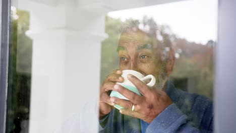 Happy-senior-biracial-man-looking-through-window-and-drinking-coffee,-unaltered,-in-slow-motion