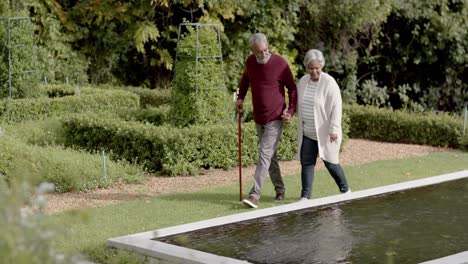 Senior-biracial-couple-holding-hands-and-walking-together-in-sunny-garden,-unaltered,-in-slow-motion