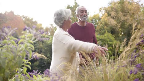 Senior-biracial-couple-walking-and-touching-flowers-in-sunny-garden,-unaltered,-in-slow-motion