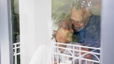 Happy-senior-biracial-couple-looking-through-window-and-embracing,-unaltered,-in-slow-motion