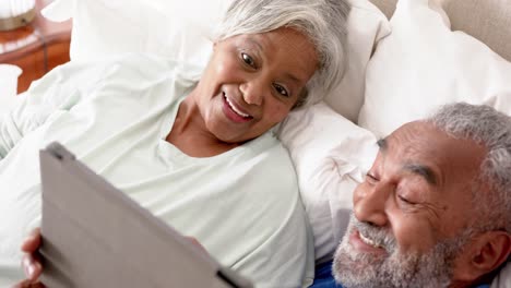 Glückliches-älteres-Gemischtrassiges-Paar,-Das-Auf-Dem-Bett-Liegt-Und-Ein-Tablet-Benutzt,-Unverändert,-In-Zeitlupe