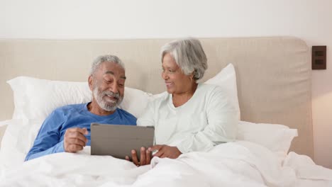 Happy-senior-biracial-couple-lying-on-bed-and-using-tablet,-unaltered,-copy-space,-in-slow-motion