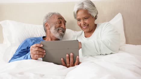 Happy-senior-biracial-couple-lying-on-bed-and-using-tablet,-unaltered,-in-slow-motion